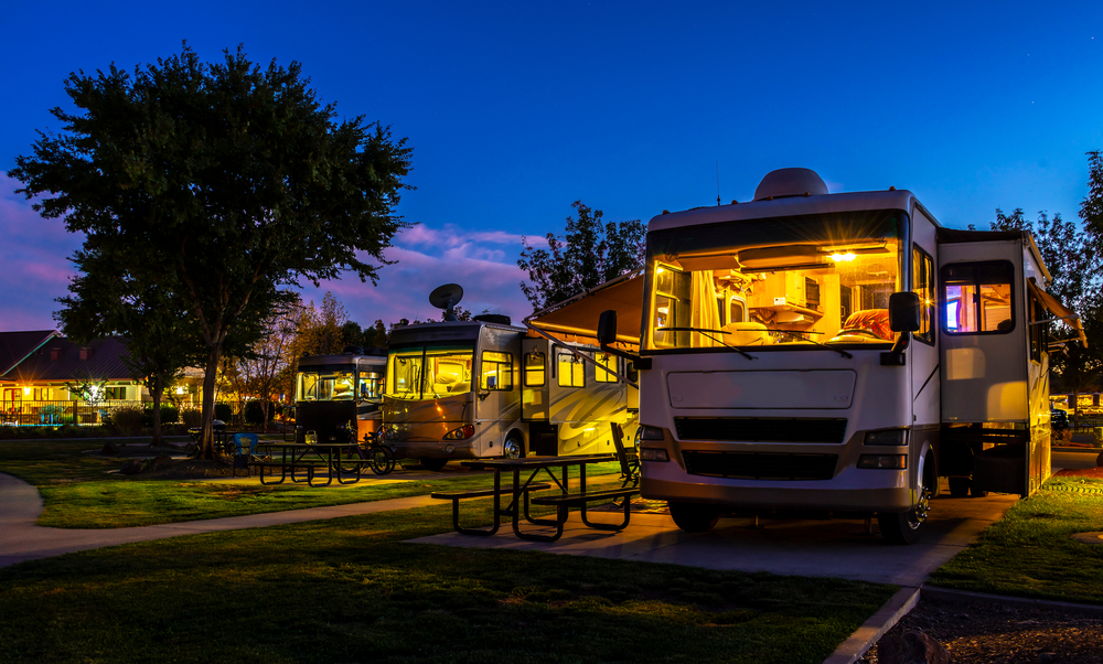 Several RVs are parked at a campsite at dusk, illuminated by warm lights inside. Picnic tables and trees are visible, and the sky is a deep blue, indicating early evening.
