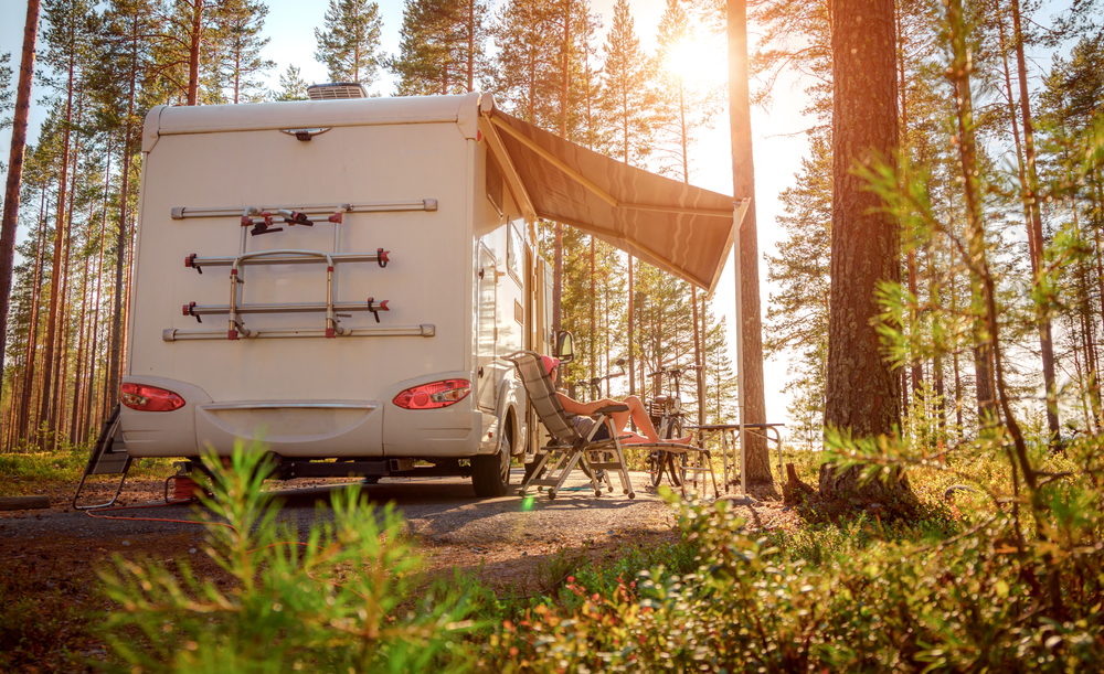 A person relaxes on a chair under the awning of a parked campervan in a sunlit forest, surrounded by tall pine trees and greenery. Camping gear is set up beside the campervan.
