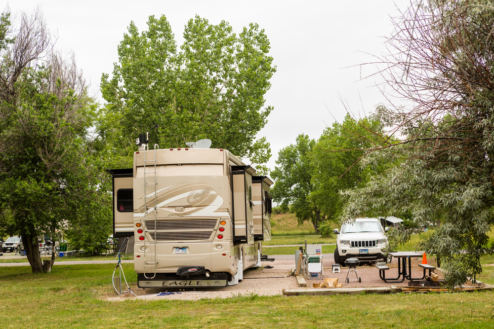 A beige RV labeled "Eagle" is parked at a campsite surrounded by trees. Nearby, a white SUV, picnic table, orange cone, and camping items are visible on a gravel pad and grassy area.